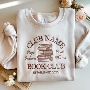 Peut inclure: Sweat-shirt crème avec texte brodé et une pile de livres. Le texte indique "CLUB NAME BOOK CLUB" avec "Page Turners" et "Book & Worms" en dessous. L'année "ESTABLISHED 2025" est en bas.