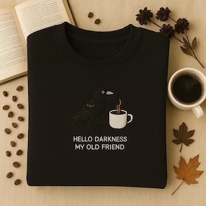 May include: Black sweatshirt with an embroidered design of a black crow and a white coffee cup with steam. The text "HELLO DARKNESS MY OLD FRIEND" is below the design. The sweatshirt is on a neutral surface.