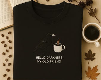 Peut inclure: Sweat-shirt noir avec un motif brodé d'un corbeau noir à côté d'une tasse à café blanche fumante. Le texte "HELLO DARKNESS MY OLD FRIEND" est en dessous. Le sweat-shirt est sur une surface neutre.