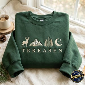 Op de afbeelding: Een donker bosgroene sweater met ronde hals voorzien van een borduursel van een hert, een bergketen, dennenbomen en een maansikkel met sterren. Het woord TERRASEN is in een lichtgekleurd lettertype onder de natuurelementen geborduurd.