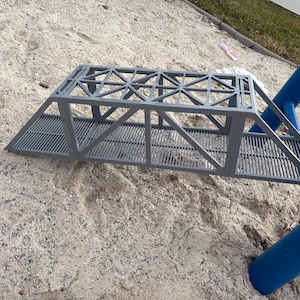 May include: A grey, 3D-printed toy bridge with a truss design, set on a sandy surface. The bridge has a slatted walkway and angled supports. A blue playground structure is visible in the background.