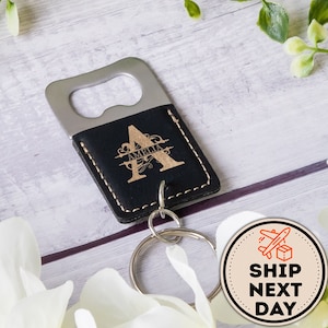 May include: A black leather bottle opener with a silver metal opener and a gold engraved initial "A" with the name "Amelia" underneath. The bottle opener is attached to a silver key ring.
