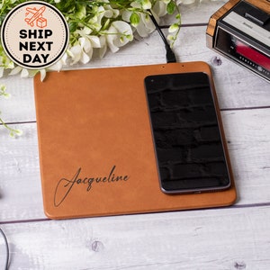 May include: A brown leather wireless charging pad with the name "Jacqueline" engraved on it. A black smartphone is charging on the pad.