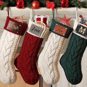 May include: Four personalised Christmas stockings with cable knit fabric. The stockings are white, red, white, and green. Each stocking has a personalised nameplate with the names Luna, Levi, Luca, and M.W.
