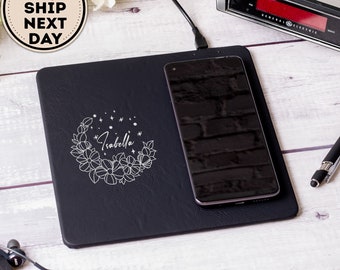 May include: Black leather wireless charging pad with a white floral design and the name "Isabella" engraved in the centre. A black smartphone is charging on the pad.