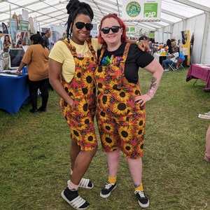 Peut inclure: Deux personnes posent pour une photo, vêtues de salopettes assorties à imprimé tournesol rouge et jaune et de lunettes de soleil. L'une porte un t-shirt jaune, l'autre un t-shirt noir. L'arrière-plan montre une tente et des personnes lors d'un événement.