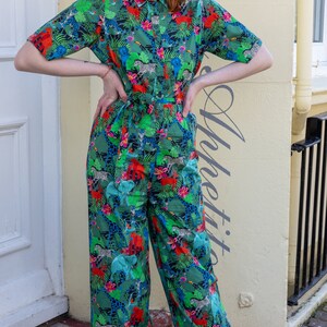 Puede incluir: Una mujer lleva un mono verde, azul y rojo con estampado de jungla que incluye animales como cebras, elefantes y tigres. El mono tiene una parte superior de estilo camisa con cuello y pantalones de pierna ancha.