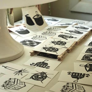 May include: A collection of small rectangular cards featuring black ink block print illustrations on off-white paper. The designs include various motifs such as a house with a heart, a beetle, a candle, a basket of strawberries, and a teapot.
