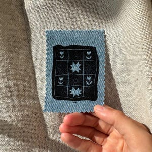 Op de afbeelding: Een rechthoekige lichtblauwe spijkerstof patch met gekartelde randen en een zwart vierkant rasterontwerp. Het raster bevat lichtblauw geborduurde motieven van vier tulpen in de hoeken en twee achtpuntige sterren in het midden.