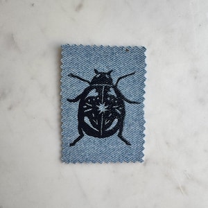 Pode incluir: Um remendo retangular de tecido jeans azul claro com bordas serrilhadas, apresentando um design de besouro bordado em preto no centro. O gráfico do inseto inclui linhas intrincadas e um pequeno detalhe em forma de estrela nas costas, criando um contraste claro contra o fundo de jeans azul texturizado.