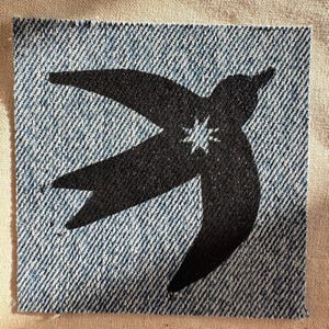 May include: A square denim patch featuring a black silhouette of a bird in flight. The bird design includes a small, unprinted star shape in the center of its body, revealing the blue and white diagonal weave of the denim fabric underneath.