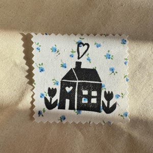 Puede incluir: Un parche de tela cuadrado con bordes dentados que presenta un fondo blanco con un pequeño patrón floral azul. Un estampado gráfico negro de una casa con una ventana en forma de corazón y una columna de humo en forma de corazón está centrado, flanqueado por dos tulipanes negros.