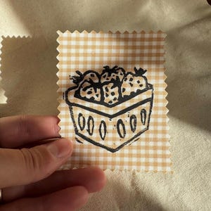 May include: A rectangular fabric swatch with pinked edges featuring a light yellow and white gingham pattern. A black ink block print of a basket filled with strawberries is centered on the fabric, showing a detailed fruit illustration.