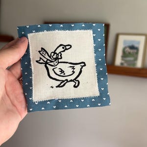 May include: A square fabric patch featuring a central cream-colored square with a black line drawing of a goose wearing a bow. The central square is bordered by a blue fabric frame decorated with a repeating pattern of small white hearts.
