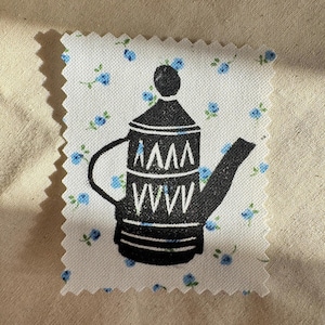 May include: A rectangular fabric swatch with pinked edges featuring a white background with a small blue floral pattern. Centered on the fabric is a black graphic print of a coffee pot with a lid, a curved handle, and a spout, decorated with white geometric triangle patterns.