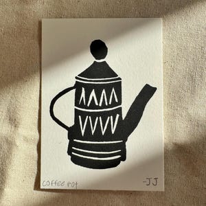 May include: A black and white block print on paper featuring a stylized coffee pot with a geometric pattern of triangles and chevrons. The text "coffee pot" is written in the bottom left corner and "-JJ" is in the bottom right corner.