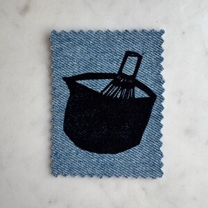 May include: A rectangular denim patch with pinked edges featuring a black screen-printed graphic of a mixing bowl and a whisk. The fabric has a visible blue twill weave texture, and the design is centered on the patch, showcasing a kitchen tool motif.