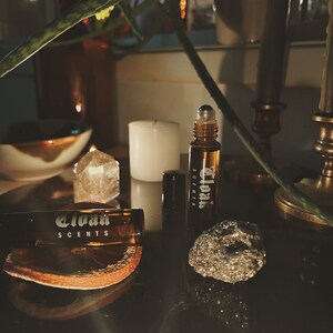 May include: Two brown glass roller bottles with the text "Eloah Scents" on the label. The bottles are sitting on a dark surface with a small, sparkly rock and a dried orange slice.