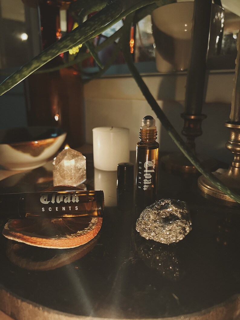 May include: Two brown glass roller bottles with the text "Eloah Scents" on the label. The bottles are sitting on a dark surface with a small, sparkly rock and a dried orange slice.