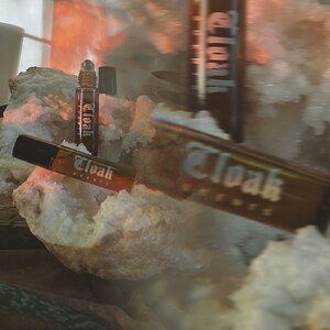 May include: Two brown glass bottles with a rollerball applicator. The bottles are labeled "Cloak Scents". The bottles are sitting on a white crystal.