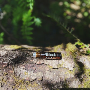 May include: A brown glass roller bottle with a white label that says "Cloak Scents" on a tree trunk covered in moss.