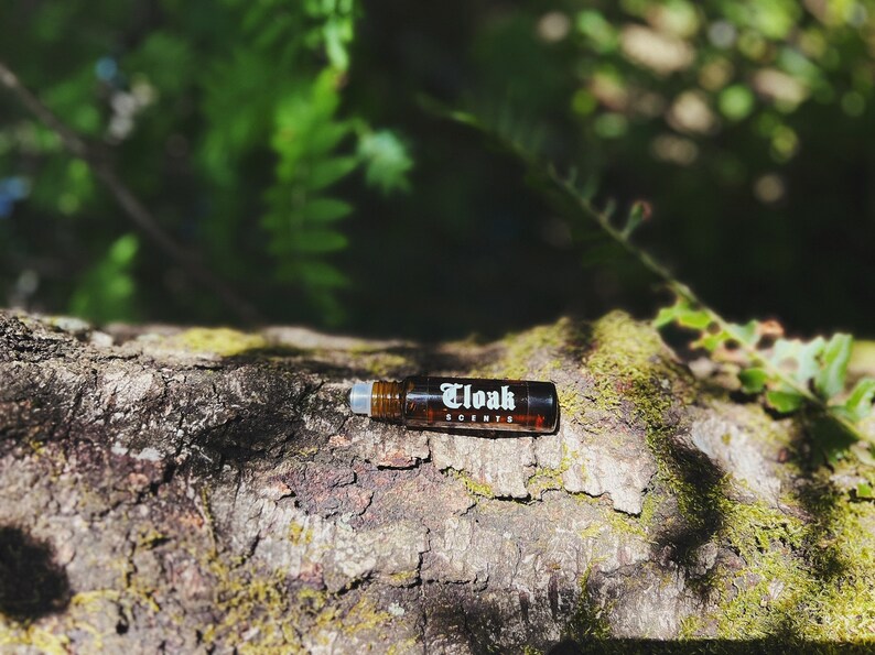May include: A brown glass roller bottle with a white label that says "Cloak Scents" on a tree trunk covered in moss.