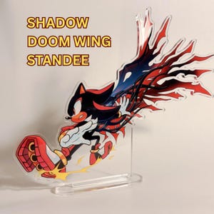 Puede incluir: Un expositor de acrílico transparente con un personaje de pelaje negro, marcas rojas y grandes alas rojas y negras. El personaje lleva calzado rojo y blanco. El texto SHADOW DOOM WING STANDEE está impreso en amarillo con un borde rojo sobre la figura.
