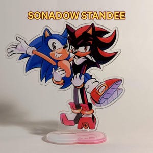 May include: A clear acrylic standee featuring a colorful illustration of the characters Sonic and Shadow. Sonic is blue with white gloves, while Shadow is black and red with white gloves. The text SONADOW STANDEE is printed above the characters.