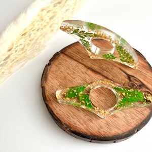 May include: Two clear resin bookmark holders with gold flecks and pressed green leaves. The bookmarks are on a wooden surface.