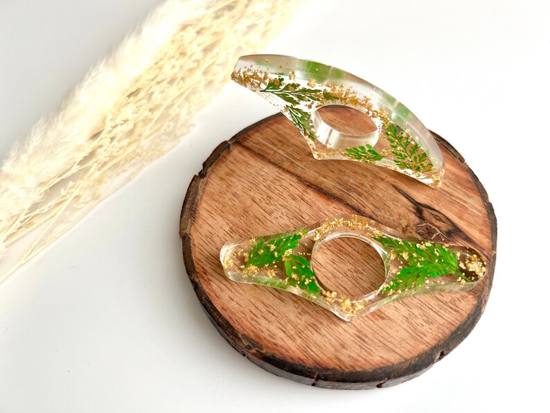 May include: Two clear resin bookmark holders with gold flecks and pressed green leaves. The bookmarks are on a wooden surface.