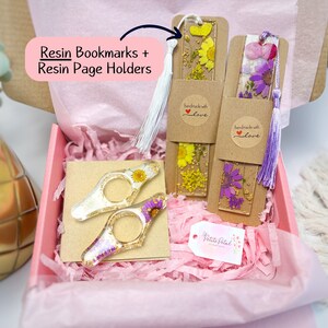 May include: A gift box filled with pink tissue paper and two handmade resin bookmarks with pressed flowers. The bookmarks have a brown paper backing with a white circle sticker that says "handmade with love". The box also contains two resin page holders with pressed flowers. The page holders are clear and have a gold accent. The box is labeled "Petite Petal Creations".