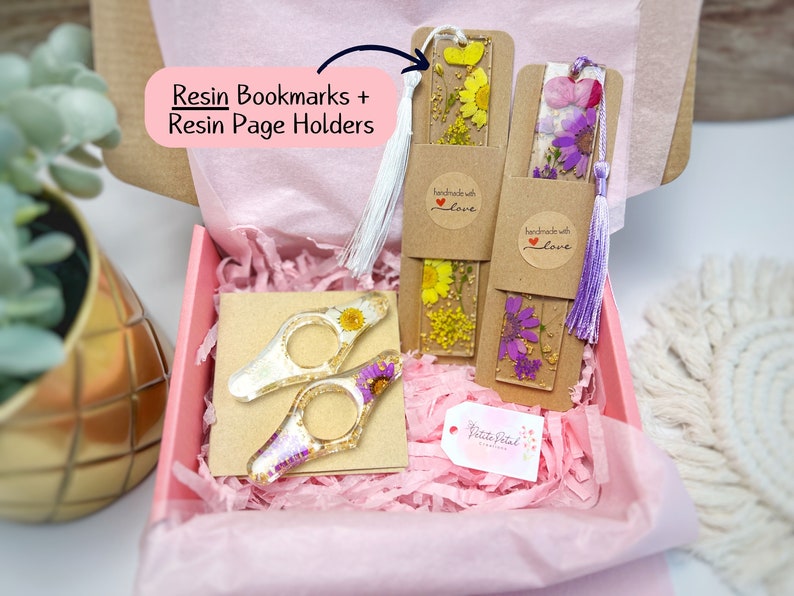 May include: A gift box filled with pink tissue paper and two handmade resin bookmarks with pressed flowers. The bookmarks have a brown paper backing with a white circle sticker that says "handmade with love". The box also contains two resin page holders with pressed flowers. The page holders are clear and have a gold accent. The box is labeled "Petite Petal Creations".