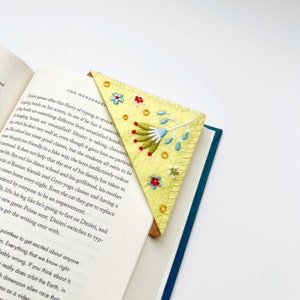May include: A yellow felt bookmark with a hand-stitched floral design. The bookmark features a green stem with blue and white flowers and red berries. The bookmark is folded into a triangle shape.