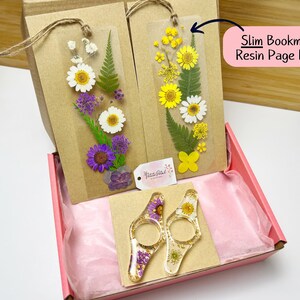 May include: Two sets of pressed flower bookmarks and two resin page holders. The bookmarks are in a clear plastic sleeve with a brown paper backing. The page holders are clear resin with pressed flowers and gold flakes. The items are packaged in a pink gift box.