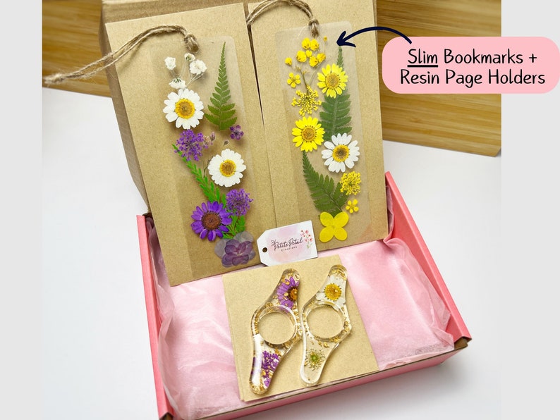 May include: Two sets of pressed flower bookmarks and two resin page holders. The bookmarks are in a clear plastic sleeve with a brown paper backing. The page holders are clear resin with pressed flowers and gold flakes. The items are packaged in a pink gift box.