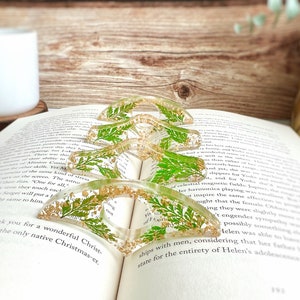 May include: Four clear resin bookmark holders with gold glitter and pressed green leaves. The bookmarks are shaped like a crescent moon and are stacked on top of each other.