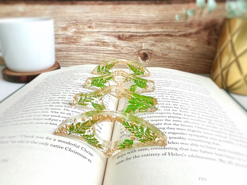 May include: Four clear resin bookmark holders with gold glitter and pressed green leaves. The bookmarks are shaped like a crescent moon and are stacked on top of each other.
