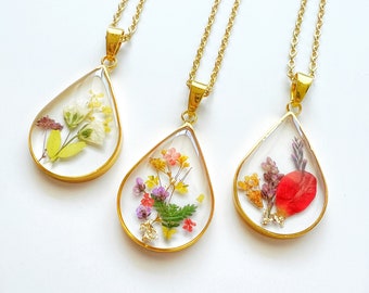 May include: Three gold necklaces with teardrop-shaped pendants filled with pressed flowers. The pendants are clear and show the flowers in detail. The flowers are yellow, pink, purple, and red.