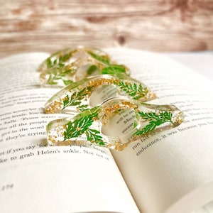 May include: Four clear resin bookmark holders with gold glitter and pressed green leaves. The text "You will be remembered" and "Much meant" is visible on the bookmarks.