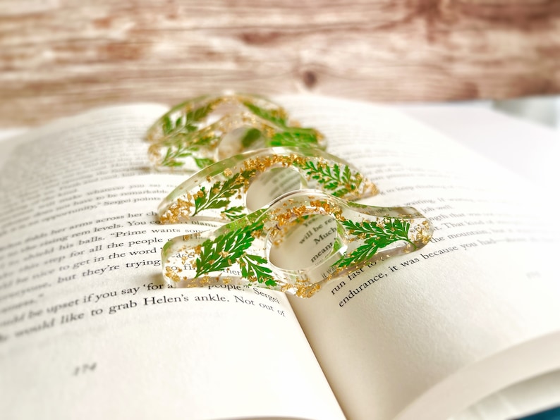 May include: Four clear resin bookmark holders with gold glitter and pressed green leaves. The text "You will be remembered" and "Much meant" is visible on the bookmarks.