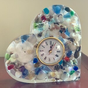 May include: A heart-shaped clock with a white face and black hands, embedded in clear resin with seashells and colorful glass beads.