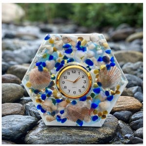 Pode incluir: Um relógio hexagonal decorativo com um mostrador dourado e algarismos pretos. O relógio é adornado com conchas, contas de vidro azuis e amarelas e pequenas pedras brancas. O relógio está sobre um leito de rochas.