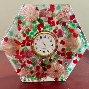 May include: A hexagonal clear resin clock with a white face and gold hands. The clock is embedded with red and green glass beads, seashells, and sea urchins.