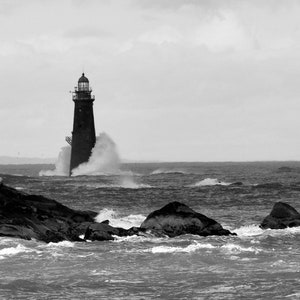 Op de afbeelding: Een zwart-witfoto van een vuurtoren die hoog boven een stormachtige zee staat. Golven beuken tegen de rotsen rond de vuurtoren en creëren een dramatisch tafereel.
