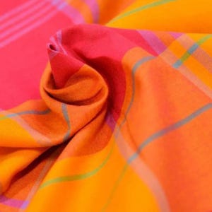 May include: A fabric featuring a vibrant plaid pattern in shades of hot pink, bright orange, and yellow. The textile is gathered in the center, creating soft folds and shadows that highlight the intersecting thin blue and purple lines.
