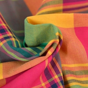 May include: A woven fabric featuring a vibrant plaid pattern in shades of hot pink, bright yellow, forest green, and deep purple. The material is folded to create soft, flowing curves that highlight the intersecting lines and colourful grid design.