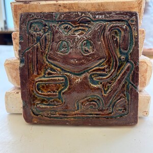 Puede incluir: Un azulejo de cerámica marrón con un diseño en relieve de un gato con ojos verdes. El gato sostiene una taza en sus patas y está rodeado de diseños abstractos.
