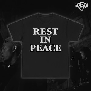 May include: Black crew neck t-shirt with the white text "REST IN PEACE" in a bold, sans-serif font. The shirt is displayed against a dark background with blurred figures on either side. A decorative emblem is in the upper right corner.
