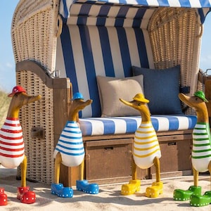 Könnte beinhalten: Vier hölzerne Entenfiguren stehen im Sand vor einem Strandkorb aus Weidengeflecht. Jede Ente trägt ein gestreiftes Hemd sowie einen passenden Hut und Schuhe in Rot, Blau, Gelb oder Grün. Der Strandkorb ist mit blau-weiß gestreiftem Stoff bezogen.