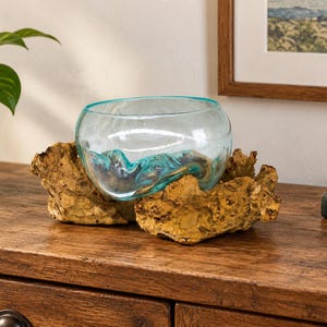 Könnte beinhalten: Eine klare, hellblaue Glasschale mit abgerundeter Form, die passgenau in die natürlichen, unregelmäßigen Konturen eines hellbraunen, strukturierten Treibholzsockels eingearbeitet ist. Das Glas hat eine glatte Oberfläche und wirkt durchscheinend.